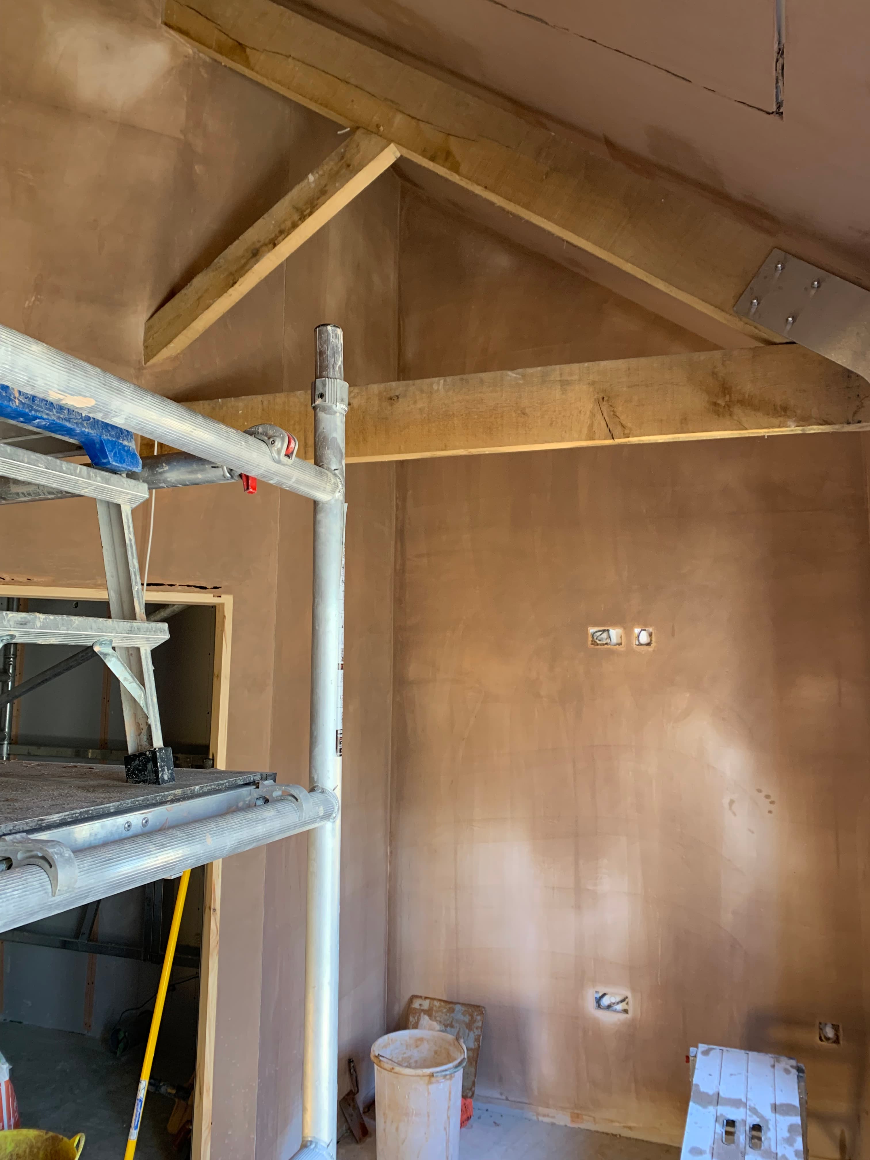 Freshly plastered room with vaulted ceiling, exposed wooden beams, and scaffolding during a home renovation.
