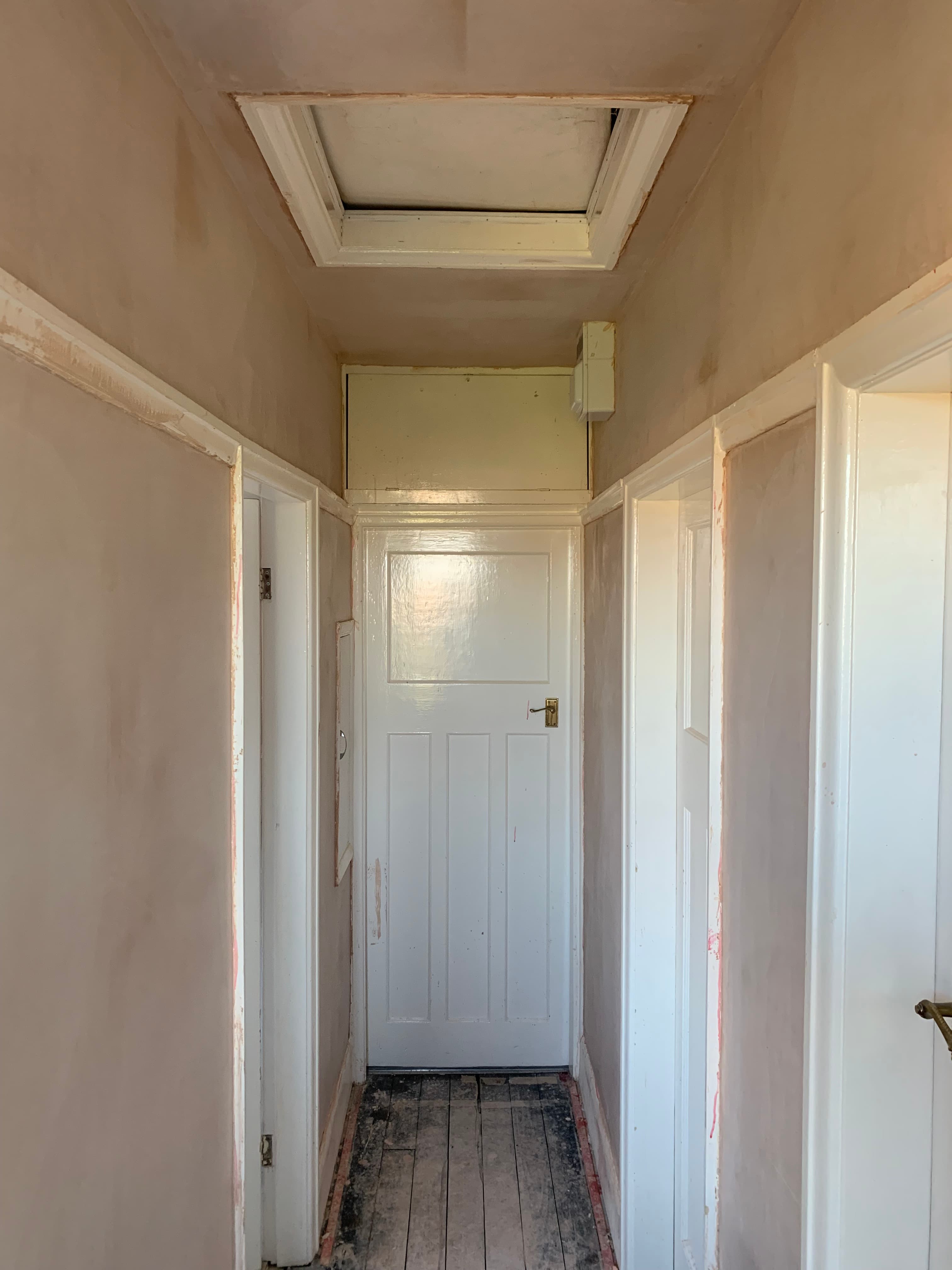 Narrow hallway with freshly plastered walls, white door, attic hatch, and exposed wooden floorboards.